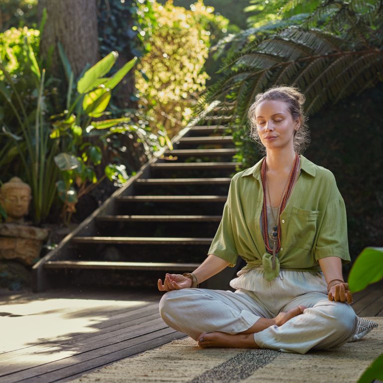 Serene woman sits in a meditative pose amidst lush greenery at a wellness retreat center, embodying mindfulness and inner peace. Practicing meditation outdoors. Calm and centered young woman embraces the healing energy of nature during a morning mindfulness session in a peaceful garden with copy space.